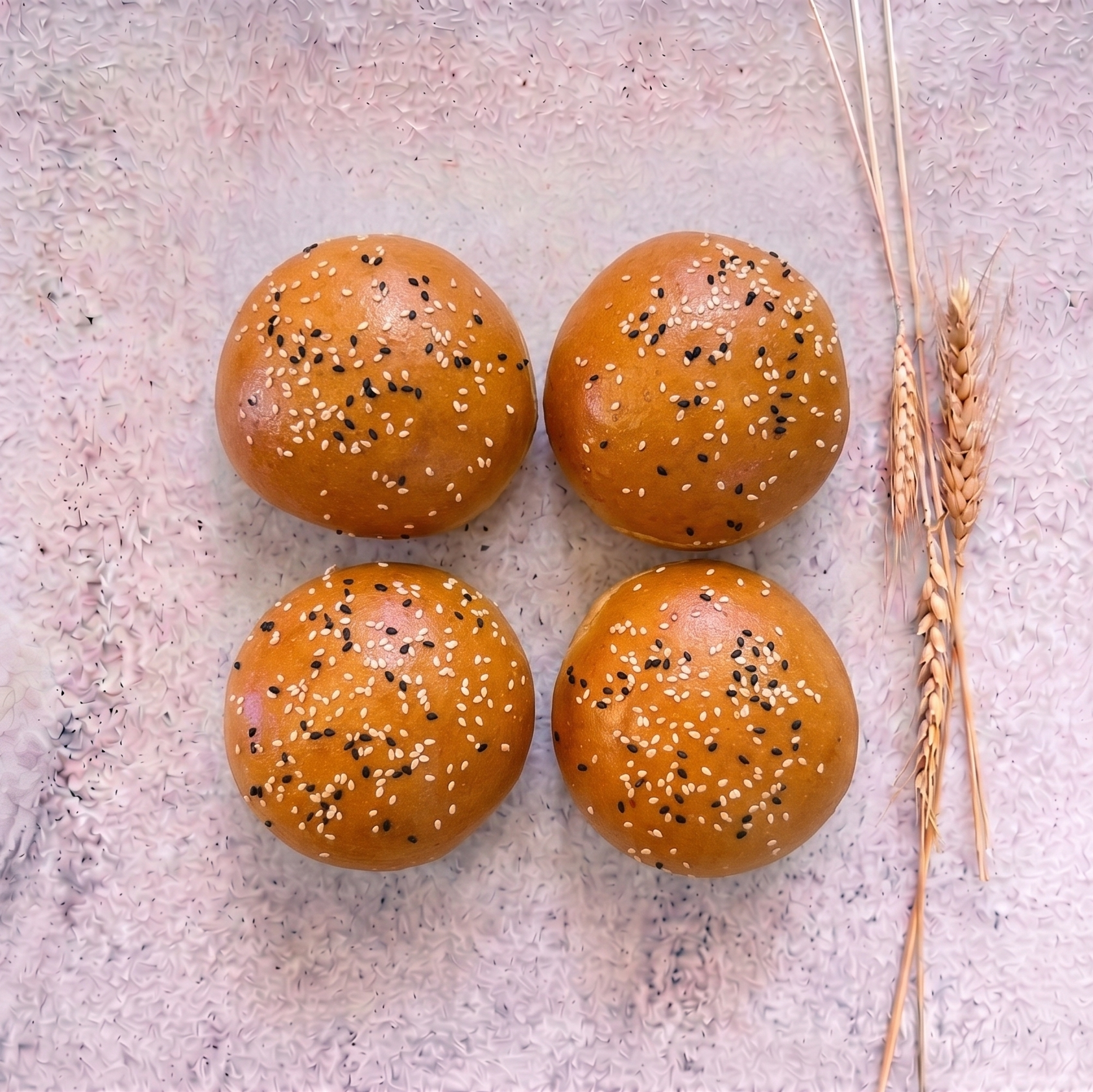 Four bread rolls with sesame seeds on a textured surface with wheat stalks.