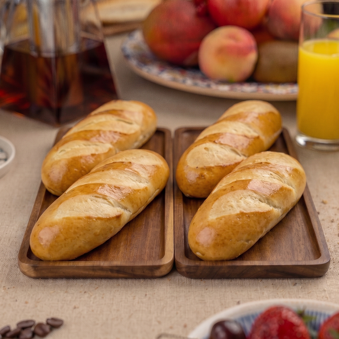 Loaves of milk bread on wooden trays with a breakfast setting including coffee, orange juice, and fruit.
